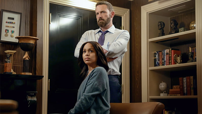 A man in a white shirt and tie stands behind a woman in a gray sweater in an office with bookshelves and medals on the wall.