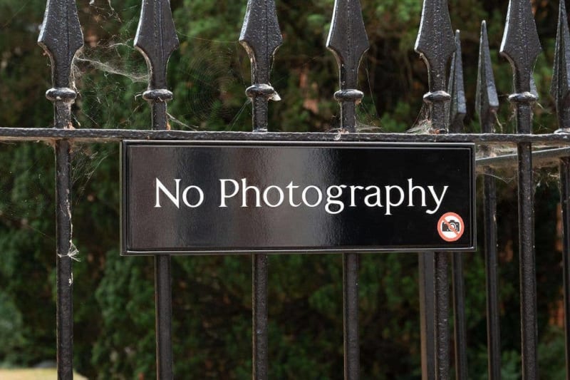 A black "No Photography" sign with a camera prohibition symbol attached to an ornate wrought iron fence.