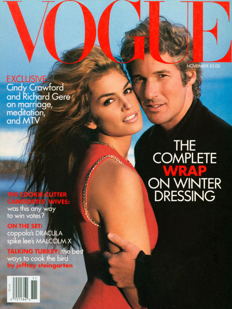 A woman with long, wind-blown brown hair wearing a red sleeveless top with a gold chain detail stands close in front of a man in a dark turtleneck sweater, both posed against a light blue outdoor backdrop on the cover of Vogue magazine.