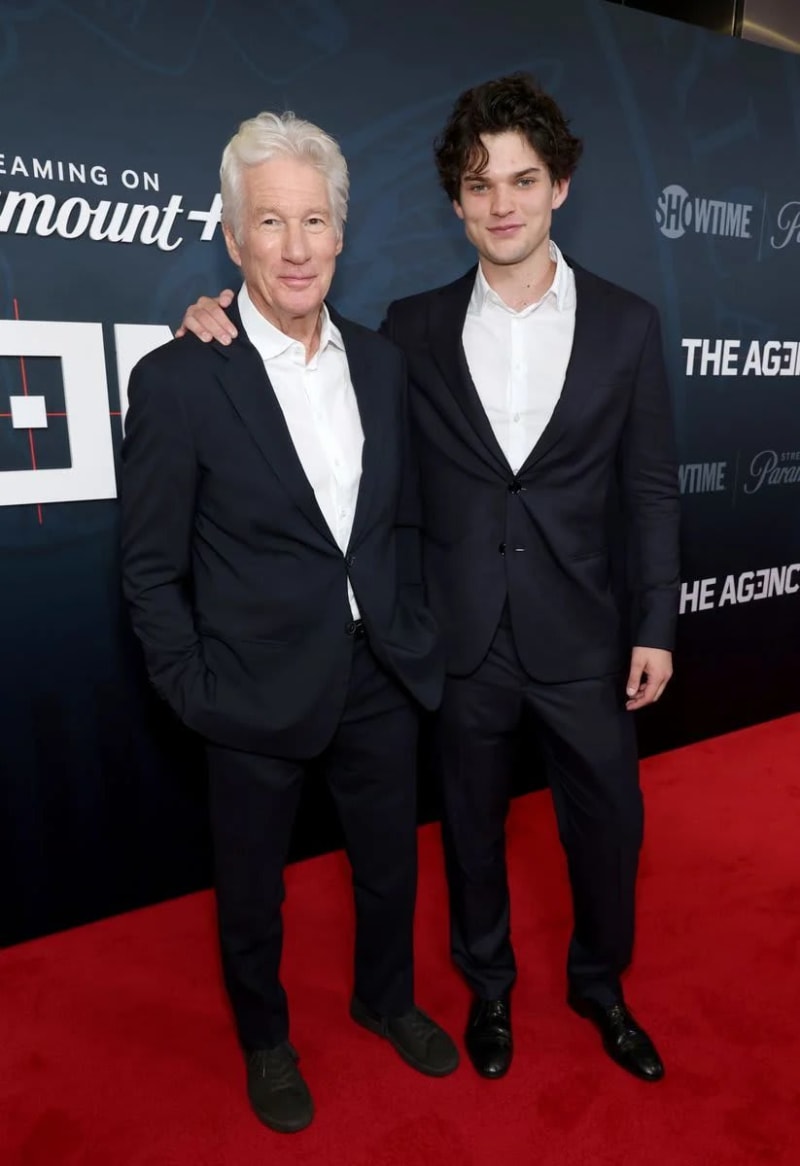 Two men in dark navy suits with white dress shirts stand together on a red carpet in front of a Showtime and Paramount+ backdrop for The Agency, with the older silver-haired man on the left and the younger dark-haired man on the right placing his hand on the other's shoulder.