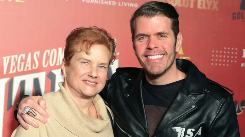 An older woman with short red hair wearing a cream turtleneck sweater stands beside a tall man in a black leather jacket, both smiling in front of a red event backdrop.