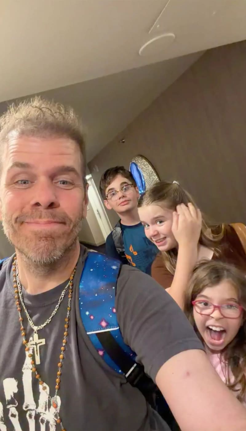 A middle-aged man with short blonde hair, a beard, a gray graphic t-shirt, and layered necklaces including a cross takes a selfie with three children in a hallway — a boy in glasses and a blue shirt, a girl with a ponytail wearing blue Stitch ears, and a younger girl with red glasses laughing with her mouth open.