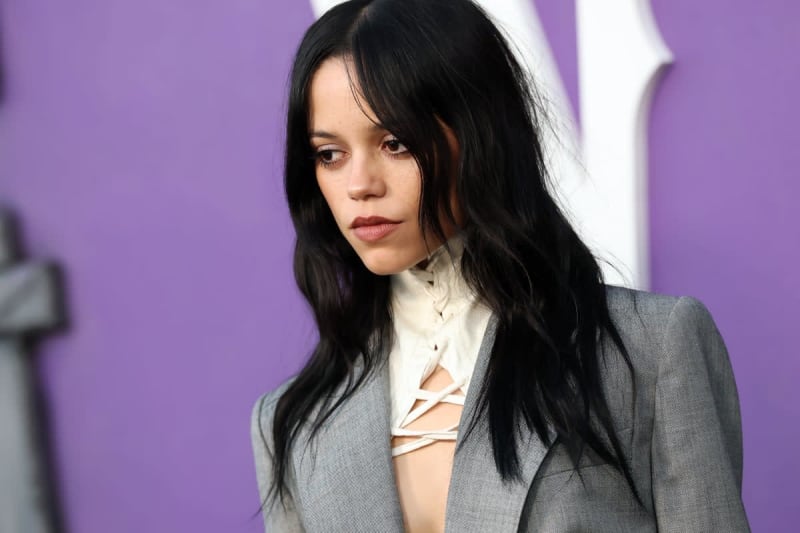 A woman with long black hair wearing a gray blazer and white lace-up top stands against a purple background.