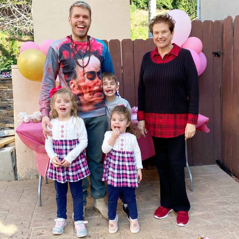 A man in a graphic sweatshirt and beaded necklace stands laughing alongside an older woman in a black sweater and red plaid shirt, with three young children in front of them, two girls wearing matching plaid skirts and a boy, all gathered on a patio decorated with pink and gold balloons.