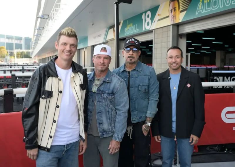 Four men standing side by side in a Formula 1 pit lane, wearing casual jackets and jeans, with Lance Stroll and Fernando Alonso garage signage visible in the background.