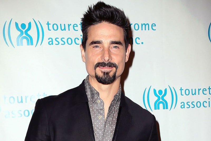 A man with dark hair, a goatee, and green eyes wearing a black blazer over a grey polka-dot shirt, posing in front of a Tourette Syndrome Association step-and-repeat banner.