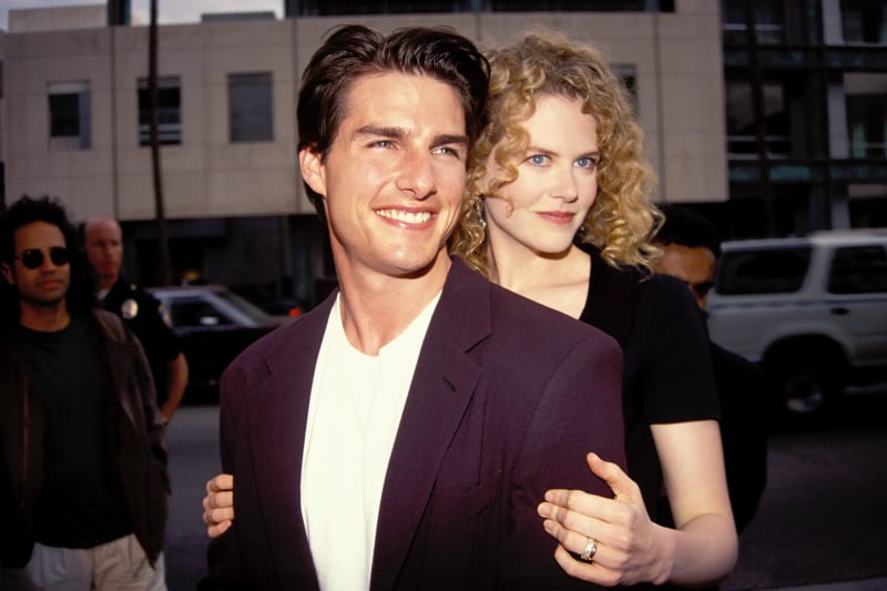 A dark-haired man in a dark purple blazer over a white shirt smiles broadly at the camera while a curly blonde-haired woman in a black short-sleeve top stands behind him with her arms around his shoulders, outdoors on a street with buildings and vehicles in the background.