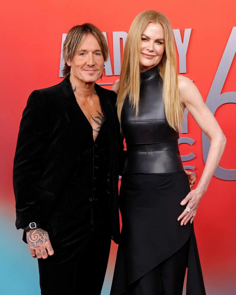 A tattooed man with medium-length streaked hair wearing an open black velvet blazer stands beside a woman with long blonde hair wearing a sleeveless black leather top and black skirt, both posing in front of a red and teal event backdrop.