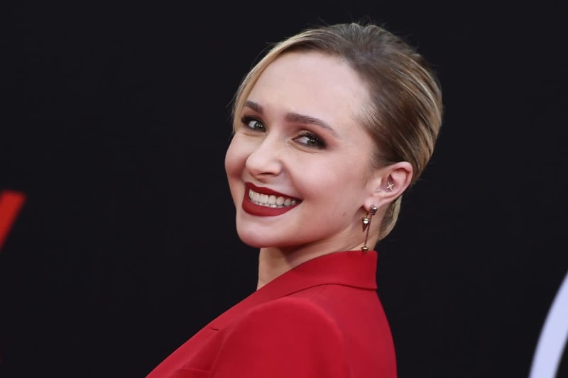 A woman with short brown hair wearing a red top and red lipstick smiles over her shoulder against a black background.