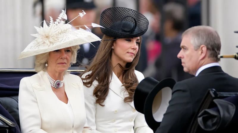 Two women in formal white outfits and elaborate hats — one in a wide-brimmed cream fascinator with feathers and a pearl-and-brooch necklace, the other in a black netted hat and double-breasted white coat — seated in an open carriage beside a man in a dark suit visible in profile on the right.