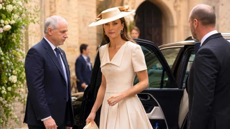 A woman in an elegant cream dress and wide-brimmed hat exits a car while two men in dark suits stand nearby at a formal event.