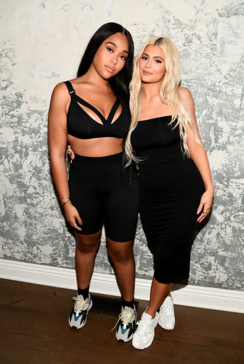 Two women posing together against a textured gray and white wall, both wearing black outfits and white sneakers.