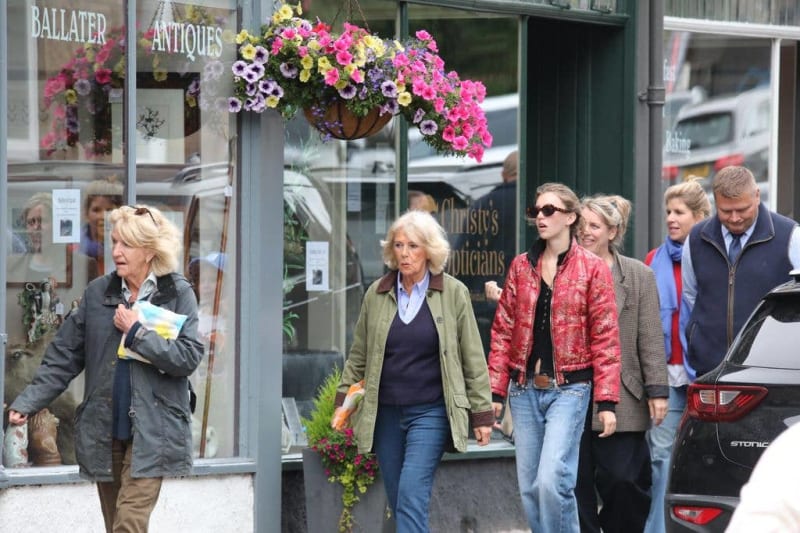 A group of people walking past an antiques shop with colorful hanging flowers, on a sunny street.