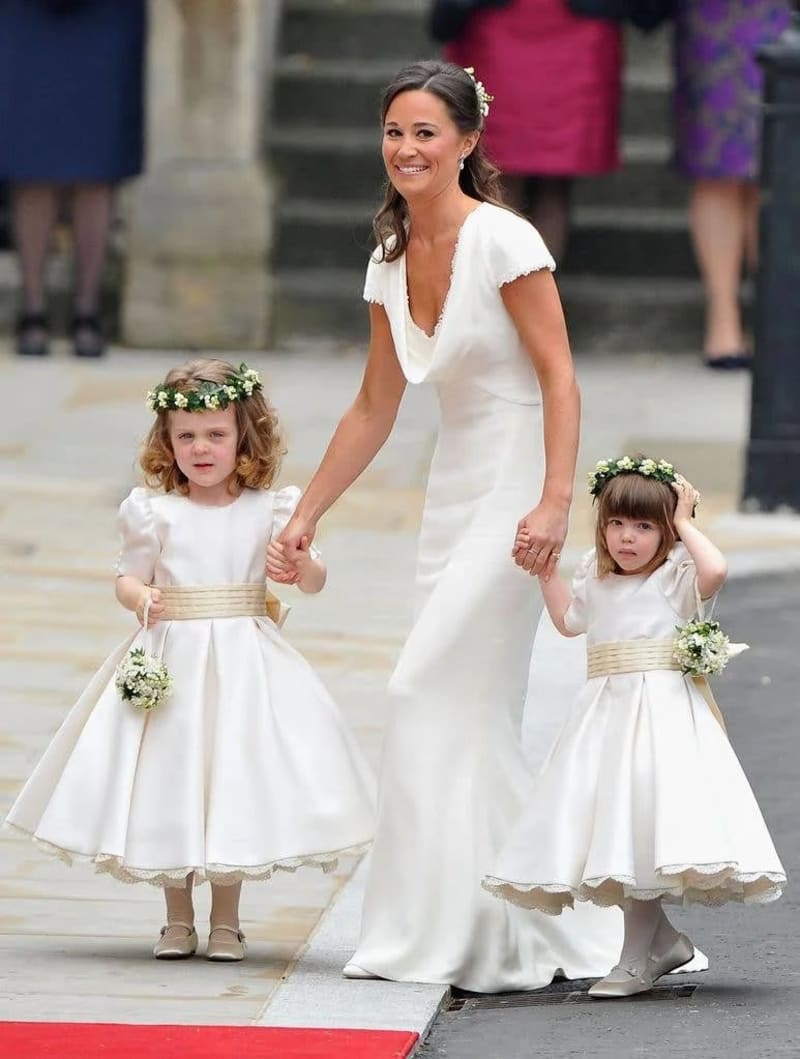 Woman in white gown holds hands with two girls in white dresses and flower crowns, walking on red carpet.