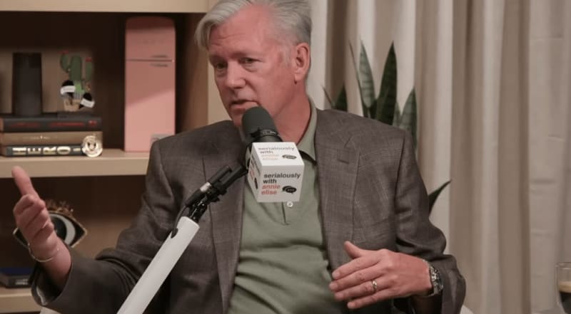 A man in a gray blazer speaks into a microphone during what appears to be a podcast or interview recording.