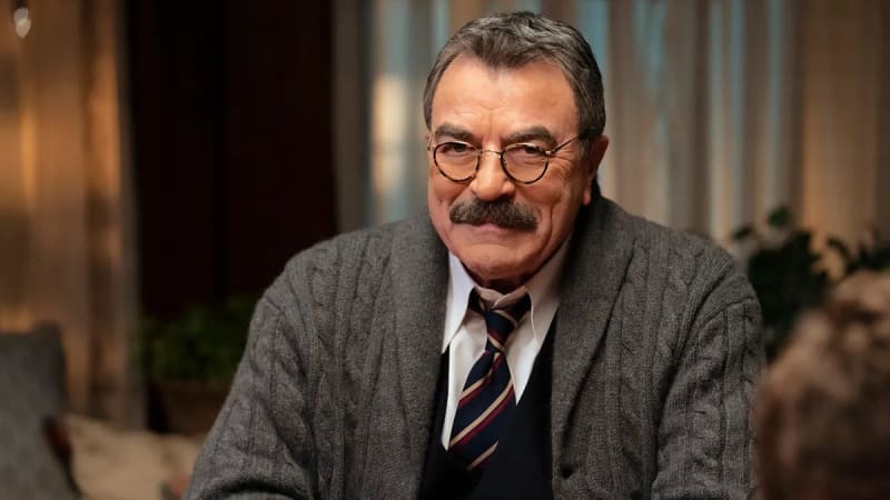 Man in gray cable-knit cardigan and striped tie, seated indoors.