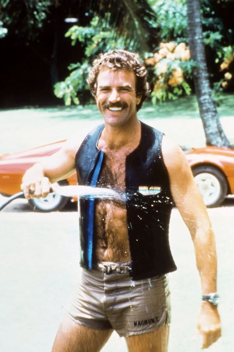 Man in open black vest and shorts, holding garden hose, red sports car and palm tree behind.
