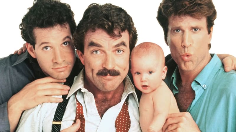 Three men posing with a baby against white background; center man has mustache and holds infant.