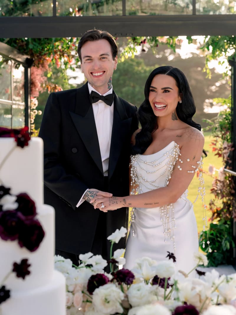 Man in black tuxedo and woman in white pearl-draped gown holding hands beside wedding cake, outdoor garden setting.