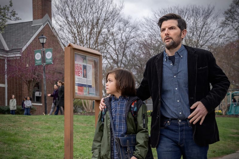 Man in black blazer and blue shirt stands behind child in green jacket near a bulletin board, outdoor school setting.