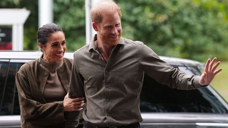Two people in casual brown clothing walk together outdoors, with one waving, near a parked vehicle and green foliage.