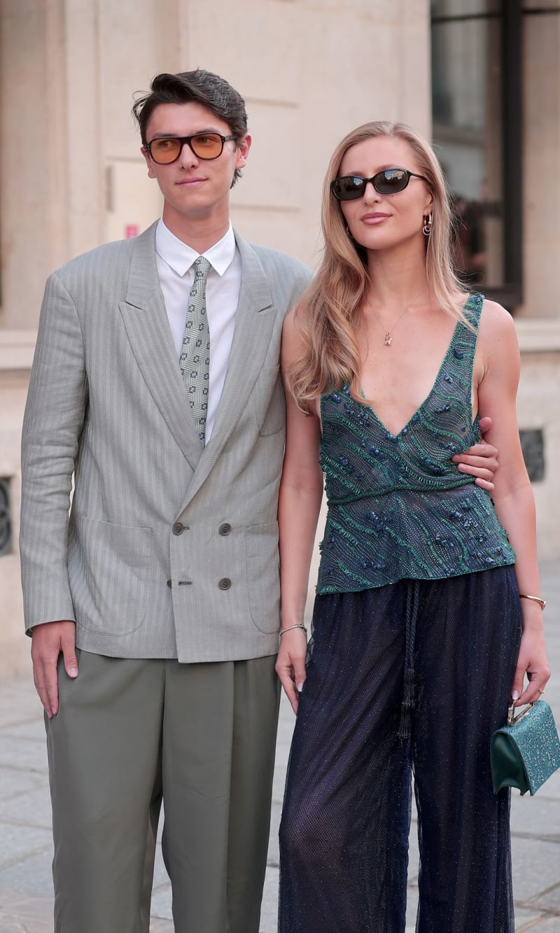 Two people posing outdoors wearing sunglasses and fashionable clothing against a neutral building backdrop.