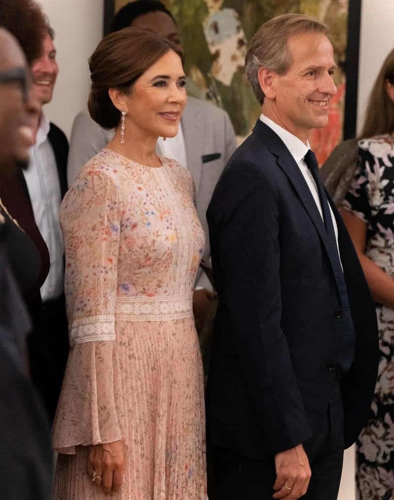 A woman in a beaded pink dress and a man in a dark suit stand together at a formal event with other guests nearby.