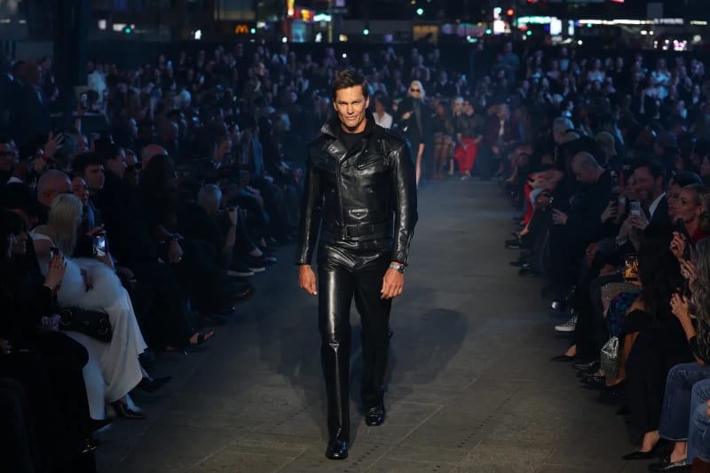 Man in all-black leather jacket and pants walking runway before crowd.