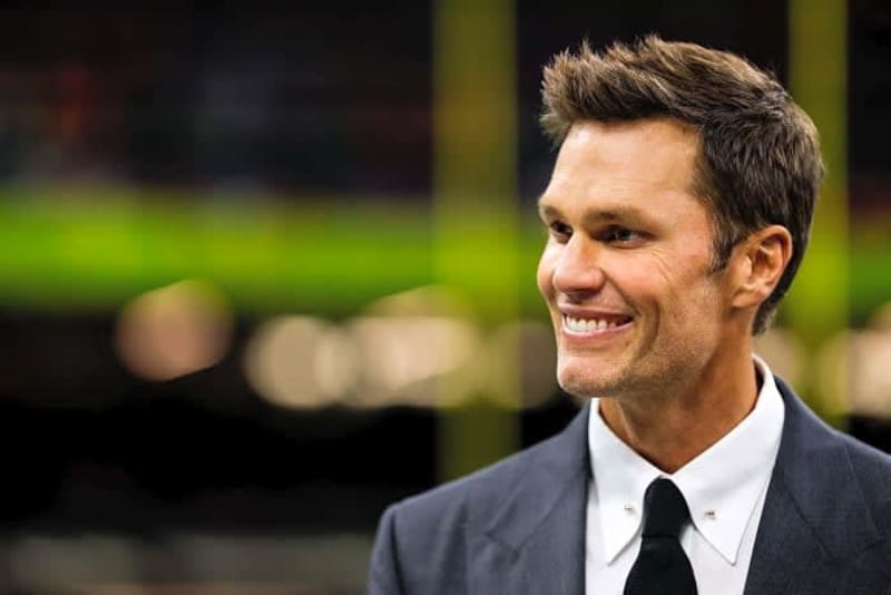 A man in a dark suit and black tie smiles while looking to the side at what appears to be a stadium or sports venue.