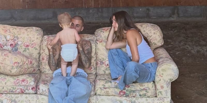 A man and woman sit on a floral couch with a toddler in a diaper standing between them in what appears to be a barn setting.