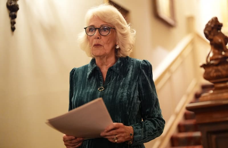 An elegant older woman with white hair and glasses wearing a dark green blouse holds a tablet in an upscale interior.
