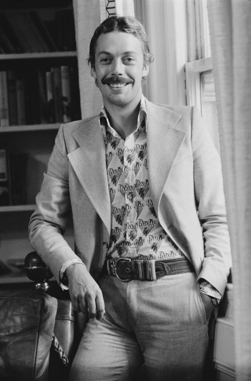 A man with a distinctive mustache sits smiling, wearing a light blazer over a patterned shirt with repeated text, in a black and white photograph.
