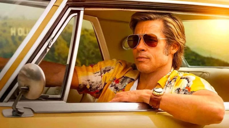 A man wearing aviator sunglasses and a colorful yellow shirt drives a vintage car with his arm resting on the window.