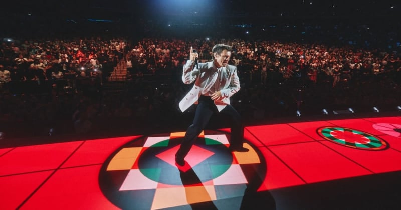 A performer in a light-colored jacket dances on a red illuminated stage floor before a large crowd in an arena.