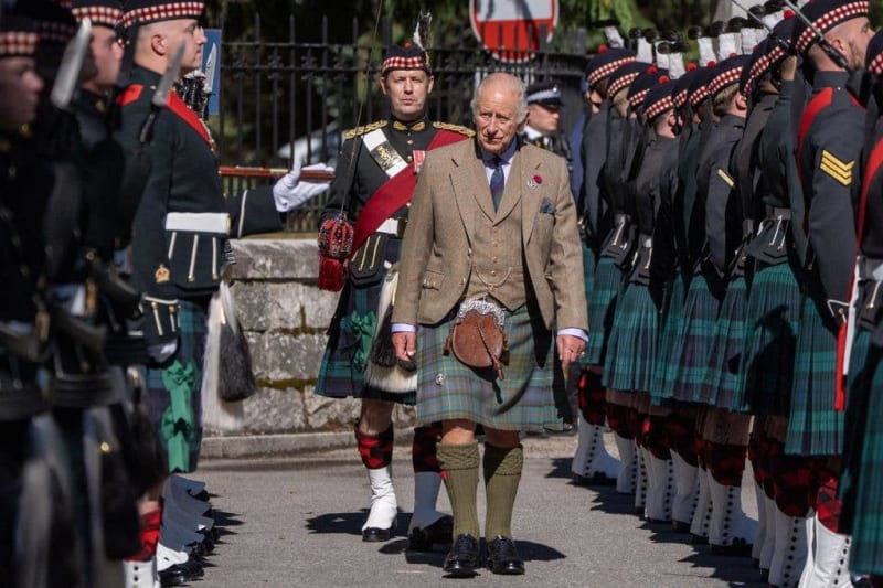 A dignitary in a tan jacket and tartan kilt walks between rows of Scottish military personnel in traditional dress uniforms.