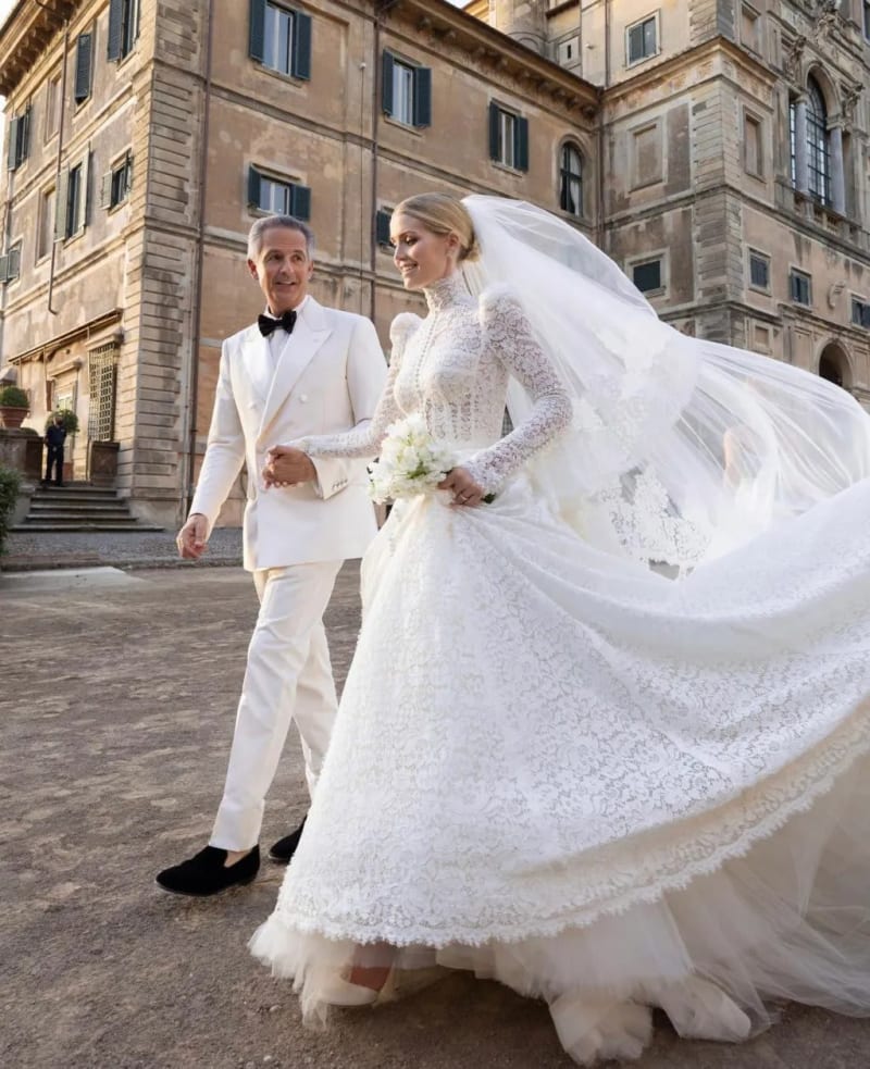 Woman in white lace ballgown with veil holding bouquet, man in white tuxedo, Italian palazzo courtyard.