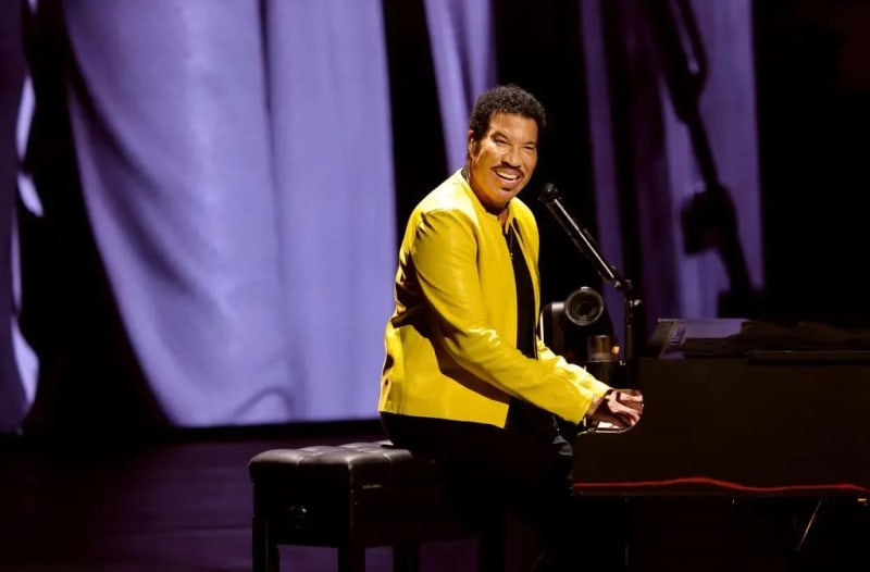 Man in yellow jacket seated at piano, smiling at microphone on stage.