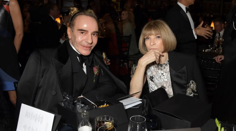 Man in black tuxedo with boutonniere and woman in floral top seated at gala dinner table.