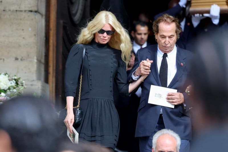 Two people in formal black attire exit a building together, with the woman wearing sunglasses and holding a clutch purse.