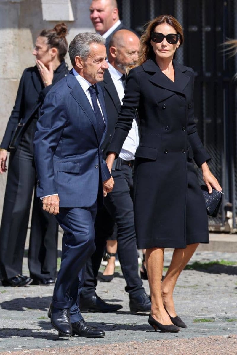 A man in a navy suit and a woman in a black coat and sunglasses walk together with security personnel in an urban setting.