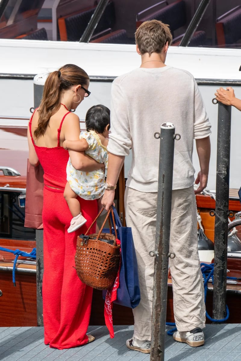 Woman in red jumpsuit holding baby beside man in white sweater, boarding a boat at a dock.