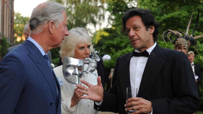 Three people in formal attire at an outdoor event, with one holding a decorative silver mask award.