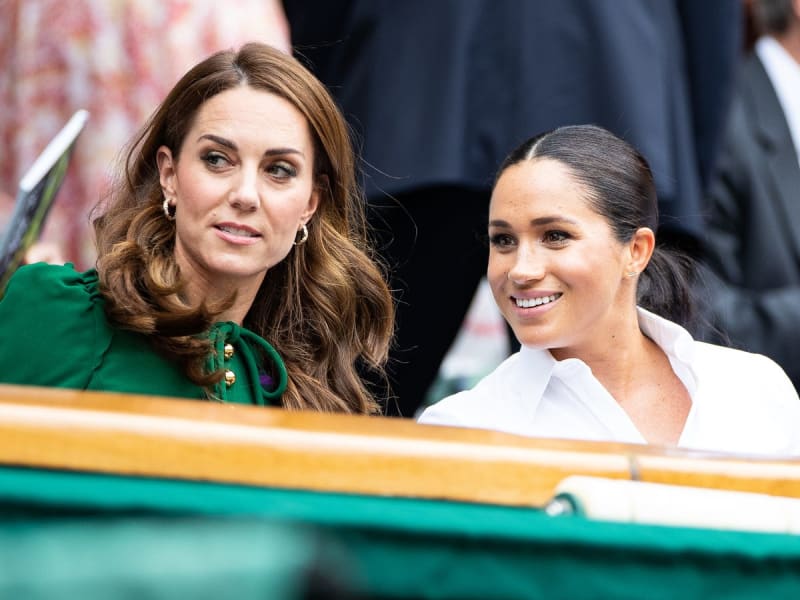 Two women seated together at what appears to be a formal outdoor event, with one wearing a green jacket and the other in white.