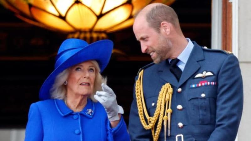 A man in RAF dress uniform with gold aiguillettes stands with a woman wearing a bright blue coat and matching hat on a balcony.