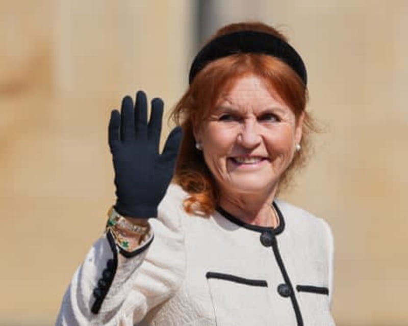A woman with reddish hair wearing a white jacket and black gloves waves while smiling at the camera.