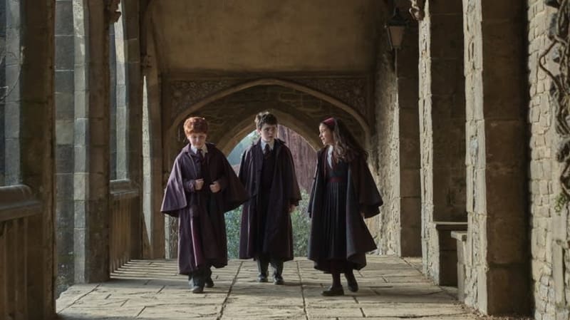 Three students in dark robes walk through a stone archway in a medieval castle corridor.