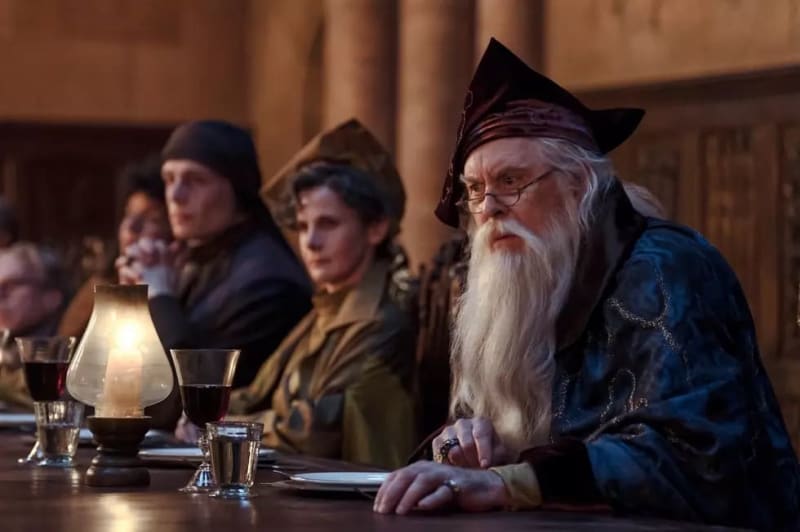 An elderly wizard with a long white beard and pointed hat sits at a table with others in a medieval hall setting.