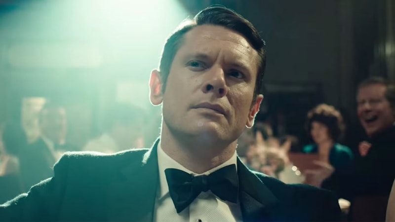Man in black tuxedo and bow tie, looking up, crowded venue behind him.