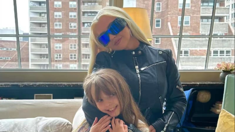 A woman wearing blue sunglasses and a black leather jacket poses with a young child in front of a window overlooking city buildings.