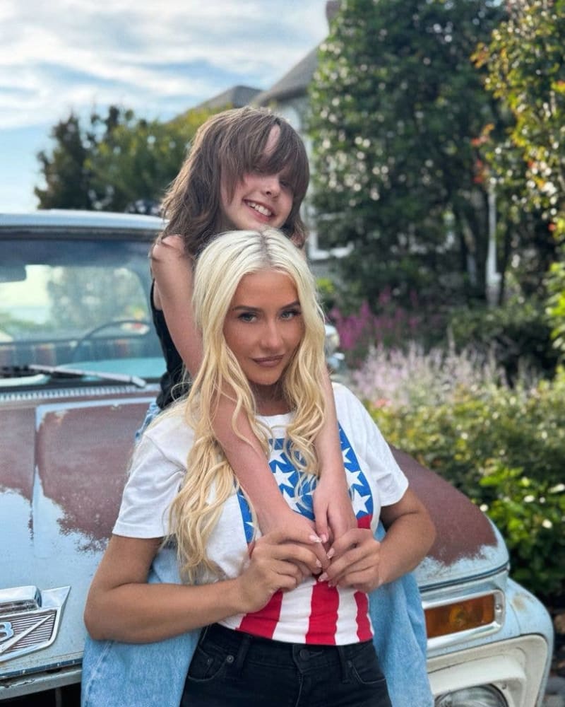 Two young women posing together outdoors near vintage cars, with lush green foliage and mountains in the background.